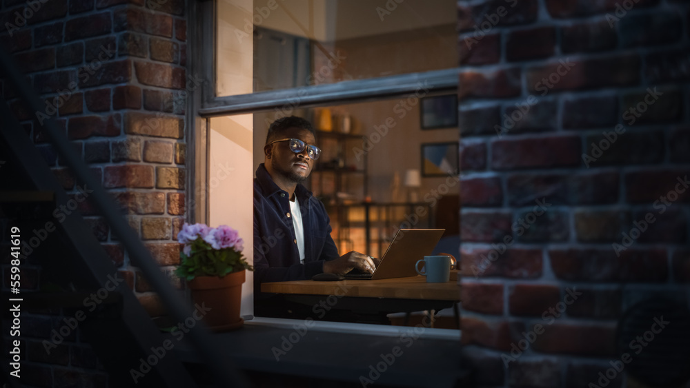 Portrait of Black Productive Young Man Working on Laptop from Home on Bedroom Desk. Businessman Developing an E-commerce Strategy and Brainstorming. He is Thinking and Typing. View Through the Window