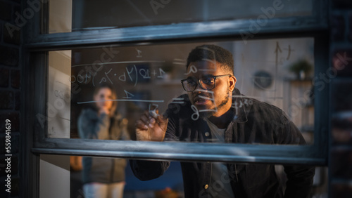 African American Young Man Using an Erasable Marker to Solve a Math Equation on Window Glass. Black Male Tutor Writing a Physics Theory and Explaining it to Fellow Female Student.