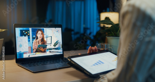 Young Asian businesswoman wear formal sit in front of desk with tablet hold stylus focus on laptop screen video online meeting with colleague in cozy living room at home night. Overtime work concept.