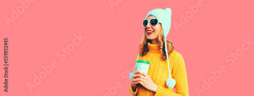 Winter portrait of happy smiling young woman with coffee cup wearing yellow knitted sweater, white hat with pom pom, heart shaped sunglasses on pink background © rohappy