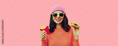 Portrait of stylish happy smiling young woman with burger and french fries fast food wearing hat on pink background