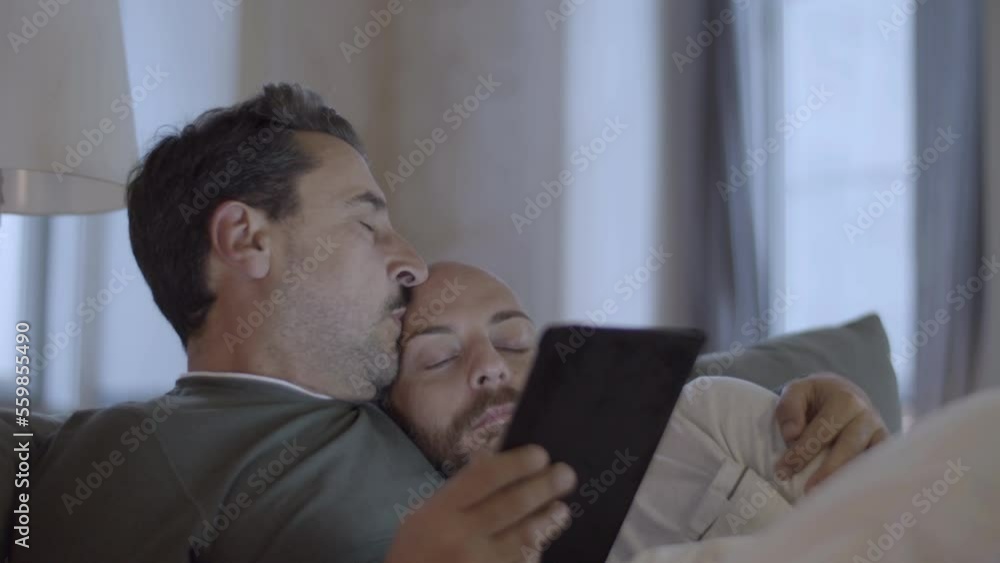 Man looking at tablet screen, lying in bed with lover in morning. Close-up shot of bearded Caucasian gay kissing his sleeping partner, relaxing on weekend together. Love, LGBT, technology concept Man looking at tablet screen, lying in bed with lover in morning. Close-up shot of bearded Caucasian gay kissing his sleeping partner, relaxing on weekend together. Love, LGBT, technology concept