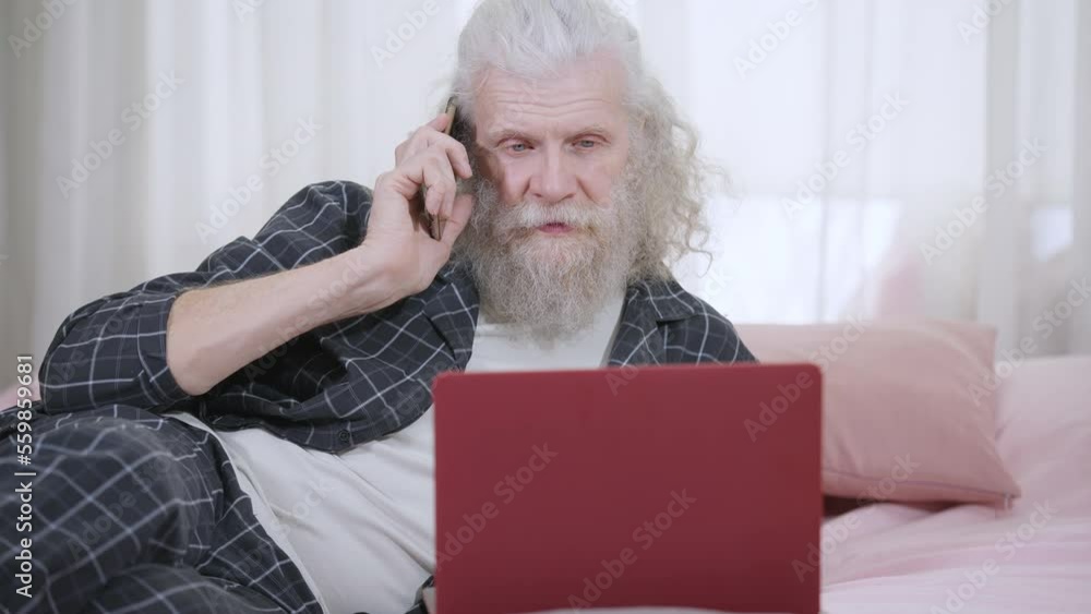 Portrait of handsome senior grey-haired man with long beard talking on phone lying on bed with laptop. Relaxed confident Caucasian retiree in pajamas enjoying morning at home in bedroom. Slow motion Portrait of handsome senior grey-haired man with long beard talking on phone lying on bed with laptop. Relaxed confident Caucasian retiree in pajamas enjoying morning at home in bedroom. Slow motion