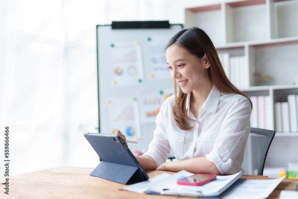 Asian business woman sitting happily with laptop and using tablet and taking intense notes on the work clipboard and smiling happily on the assignment. Asian business woman sitting happily with laptop and using tablet and taking intense notes on the work clipboard and smiling happily on the assignment.