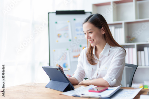 Asian business woman sitting happily with laptop and using tablet and taking intense notes on the work clipboard and smiling happily on the assignment.