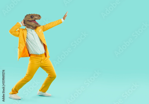 Strange guy in reptile costume dancing and having fun. Crazy young man wearing yellow suit and funny dinosaur mask dancing and moving hands isolated on blank turquoise copy space background