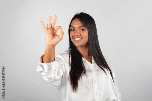 Cheerful millennial black lady in white blouse show ok sign with hand, isolated on gray background