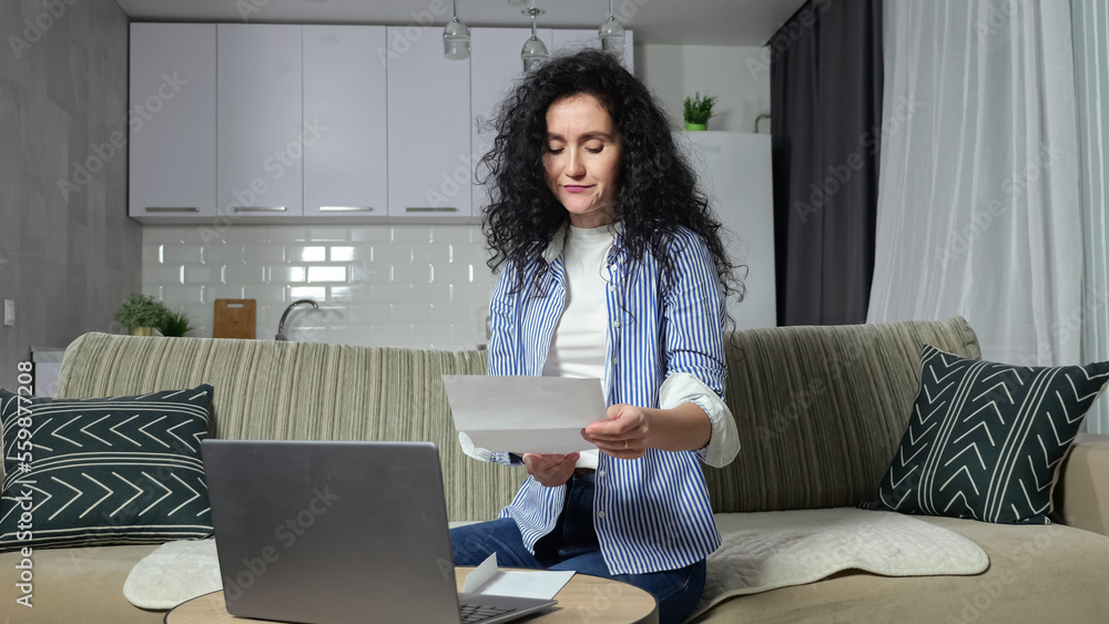 Adult woman checks utility bills and enters information into laptop. Black-haired housewife looks at bills and tries to pay communal payments via Internet from home