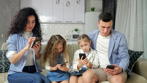 Family sits on sofa looking at smartphones and explores Internet at home. Addicted parents and girl siblings look at phones with zombified expressions