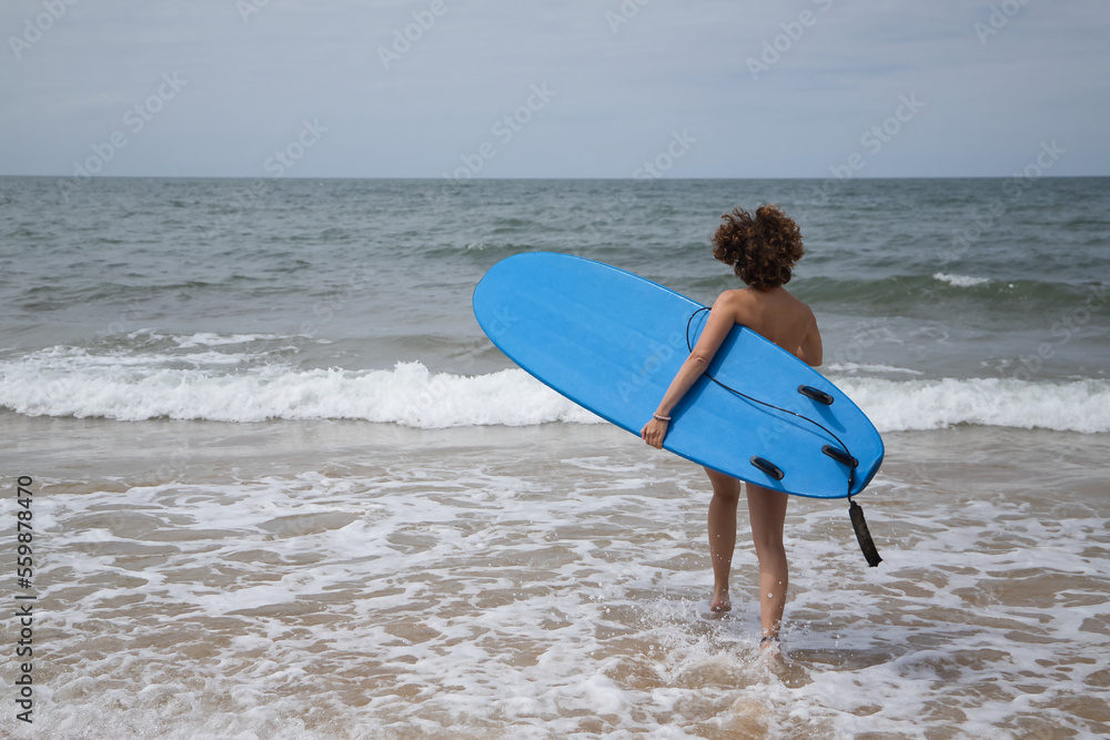 Attractive mature woman with curly hair, sunglasses and bikini, running towards the water holding a blue surfboard under her arm. Concept sea, sand, sun, beach, vacation, surf, summer.