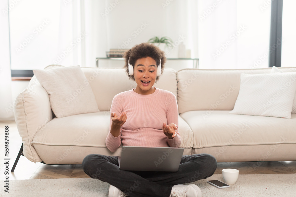Happy surprised young mixed race woman in wireless headphones looking at laptop, talking, has video call Happy surprised young mixed race woman in wireless headphones looking at laptop, talking, has video call