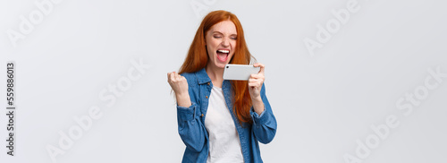 Confident and sassy good-looking redhead woman beat score, watching tennis match online on smartphone and cheering for player, fist pump delighted, celebrating success, winning in mobile game photo