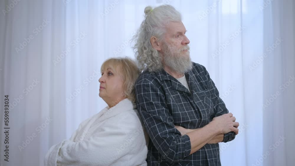 Argued senior couple standing back to back with crossed hands thinking looking away. Medium shot portrait of sad Caucasian husband and wife in pajamas at home after quarrel. Slow motion Argued senior couple standing back to back with crossed hands thinking looking away. Medium shot portrait of sad Caucasian husband and wife in pajamas at home after quarrel. Slow motion