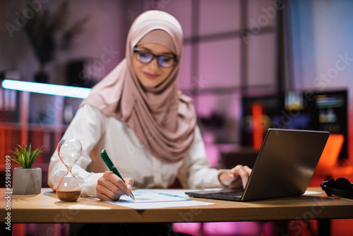 Focus in hand with pen. Attractive confident muslim business woman, office manager, wearing hijab using laptop while making financial report while writing on paper working at night.