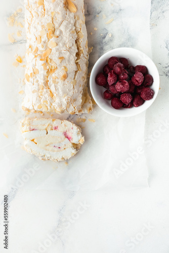 Photos Cut of homemade meringue roll decorated with almond flakes and freeze-dried red berries on the white marble background