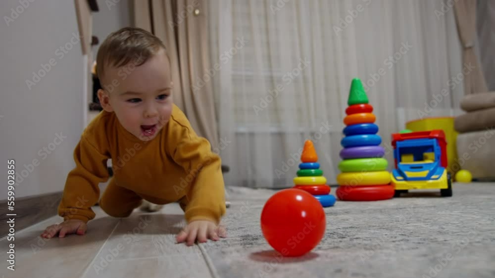 Active Caucasian toddler wearing orange clothes crawls around the room among the toys. Baby follows the red balls kicking it by the floor.