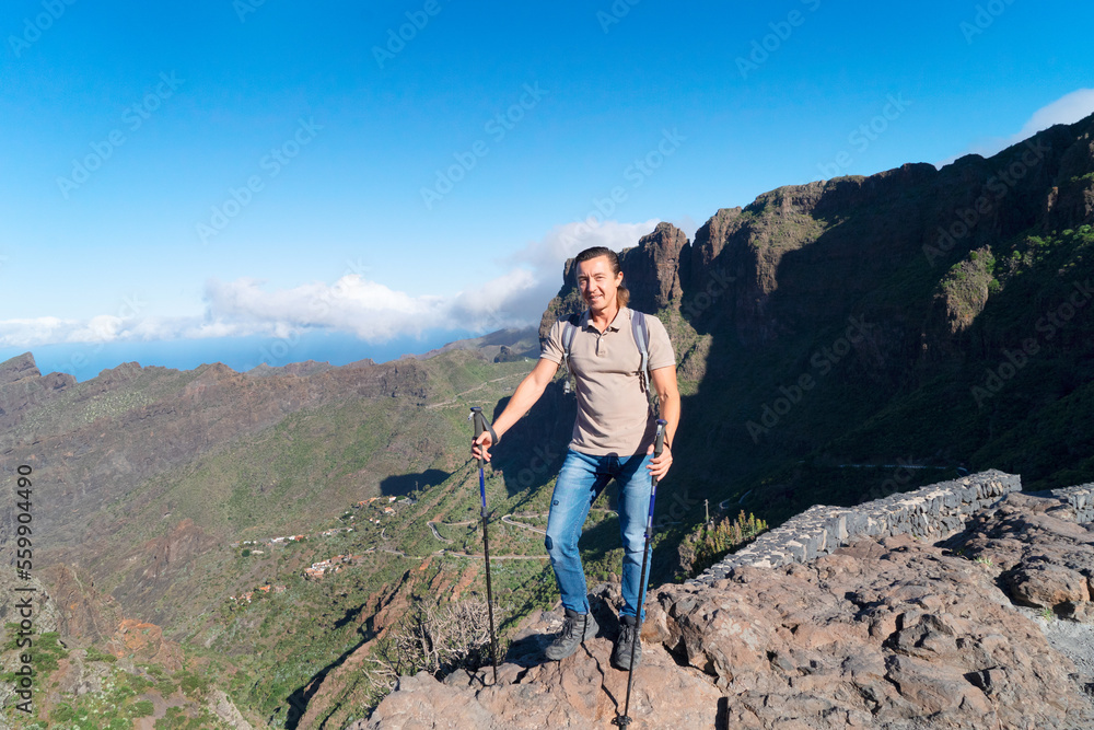 Happy middle aged man traveler smiling at camera while standing on top of hill against blue sky and beautiful landscape, hiking in mountains and enjoying adventure. Active lifestyle concept