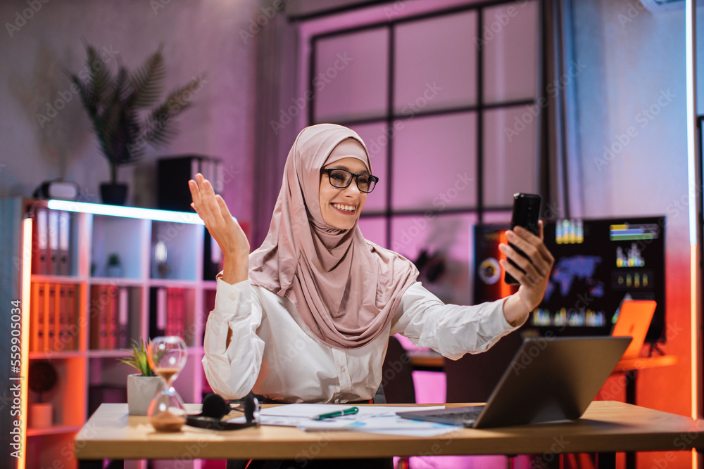 Portrait of smiling muslim businesswoman having mobile video conversation while sitting at table and working on wireless laptop. Working process at office of young girl at evening time. Portrait of smiling muslim businesswoman having mobile video conversation while sitting at table and working on wireless laptop. Working process at office of young girl at evening time.