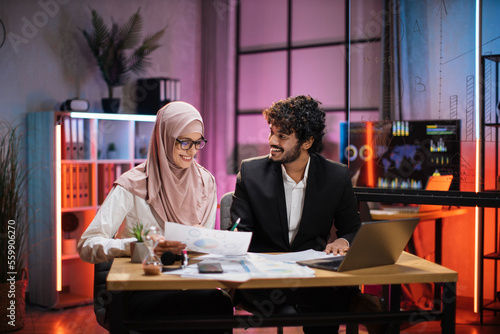 Fényképezés Two multicultural company workers male financial expert and muslim female office worker using laptop, showing paper report with graphs and charts during video meeting with colleagues in office