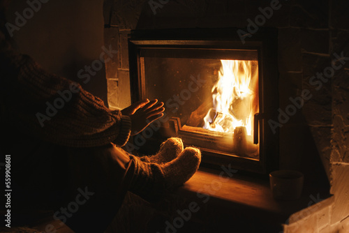 Stylish woman warming up hands and feet at cozy fireplace in evening. Fireplace heating in house, electricity blackout. Atmospheric time at fireside in home