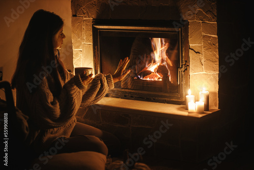 Woman hands holding cup of tea and warming up at cozy fireplace in dark evening room, close up. Fireplace heating in house, electricity blackout. Atmospheric time at fireside in home © sonyachny