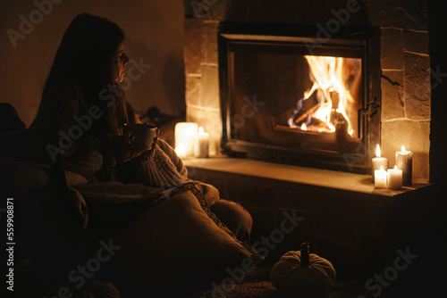 Obraz na płótnie Stylish woman with warm cup of tea sitting at cozy fireplace in dark evening room