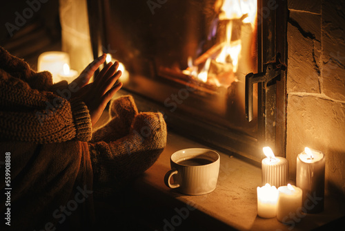 Fototapeta Stylish woman warming up hands and feet at cozy fireplace with tea in evening