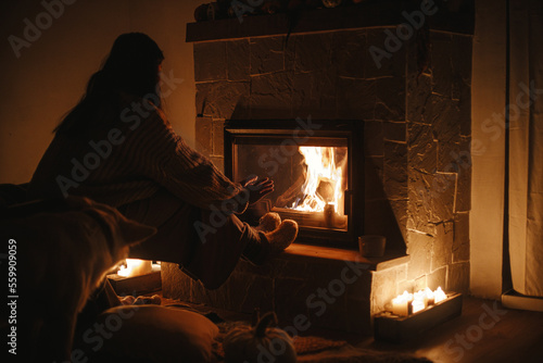 Stylish woman warming up hands and feet at cozy fireplace in evening. Fireplace heating in house, electricity blackout. Atmospheric time at fireside in home © sonyachny