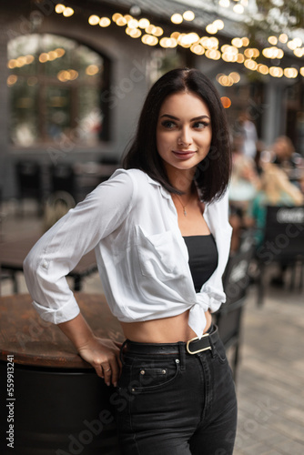 Fashion beautiful woman with stylish black jeans and white shirt stands in the city