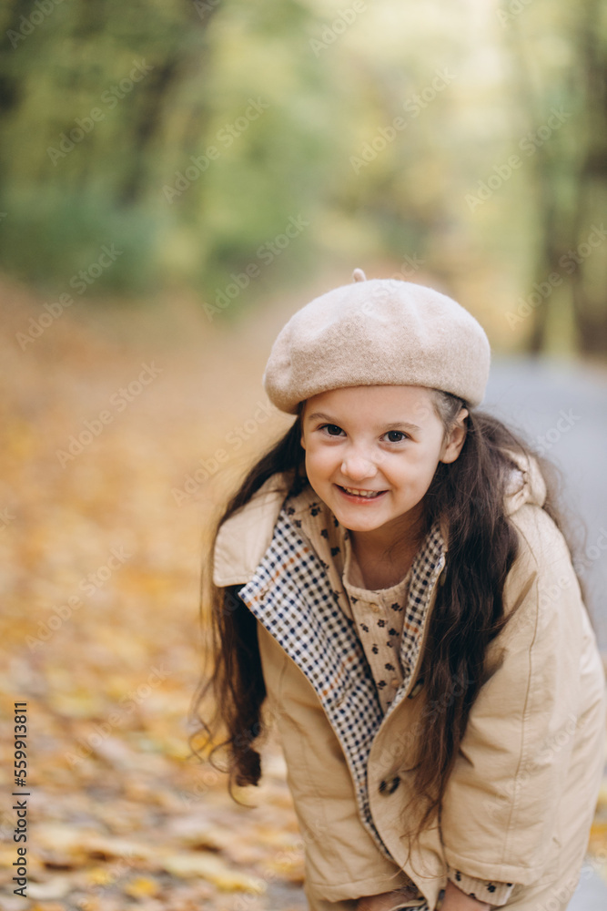 Portrait of happy little girl in beige coat and beret holding yellow maple leaves and spending time in autumn park