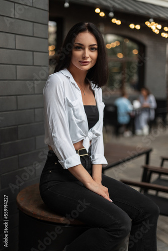 Fashionable beautiful urban business girl in stylish clothes with a white shirt, black jeans and top sits on a black barrel in the city with lights bokeh