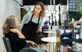 Portrait of young woman professional hair stylist talking to elderly female client in salon, choosing new hairdo .