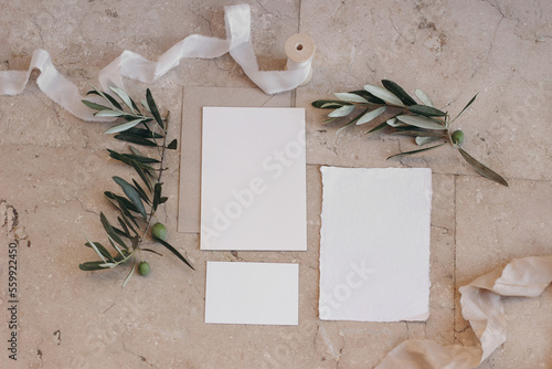 Summer wedding stationery set, suite. Italian, Greek mock-up scene with blank paper greeting cards, envelope on beige marble tiles background. Green olive tree branches, silk ribbon. Flat lay, top.