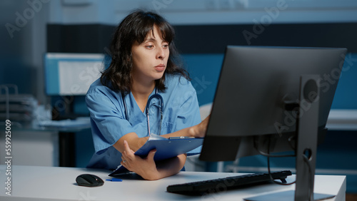 Physician nurse checking illness report on computer while working at patient treatment to help cure disease during night shift in hospital office. Medical assistant looking at papers with expertise