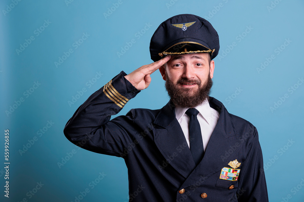 Smiling airplane captain saluting, wearing aviation uniform and hat front view portrait, plane captain looking at camera. Aviation academy pilot with airline wings badge on jacket Smiling airplane captain saluting, wearing aviation uniform and hat front view portrait, plane captain looking at camera. Aviation academy pilot with airline wings badge on jacket