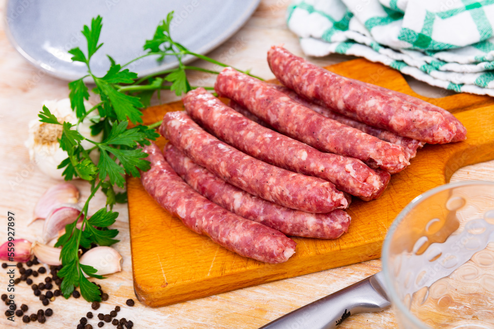 Fresh raw pork longaniza sausages ready for grilling on wooden cutting board with kitchen knife, condiments and greens. Spanish cuisine