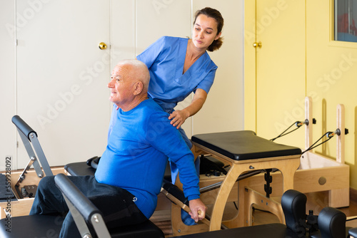 Personal female trainer controlling movements of mature man doing pilates on reformer in fitness studio.