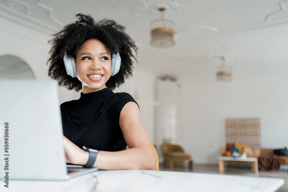 A happy young woman smiling is sitting at her workplace and using her laptop and headphones to have a video chat with someone at home.
