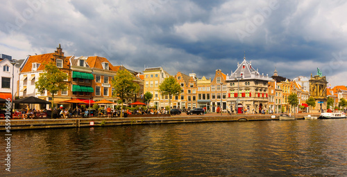 View of riverside of Spaarne with moored boats on water. Cloudy townscape of Haarlem, province of North Holland, Netherlands.