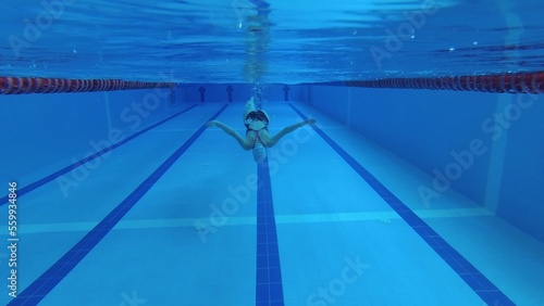 sportswoman swimmer in a tracksuit, in diving goggles, in a swimming cap is training in the pool. The athlete swims underwater. Underwater shooting, wide angle, 4k.