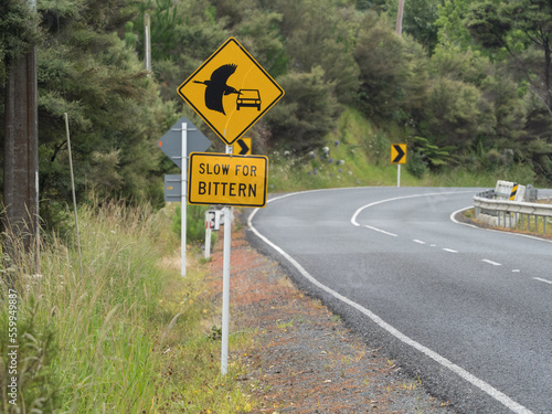 A rectangular roadside sign says 'slow for bittern'. A black silhouetted picture of a flying bittern and car can be seen above on a diamond shaped sign. Yellow signs.Road in background.