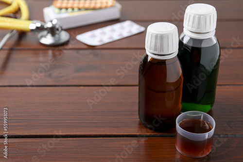 Bottles of syrup and measuring cup on wooden table, space for text. Cold medicine