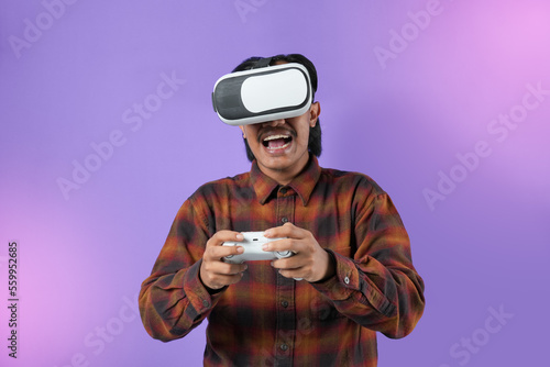 Expression of amazed asian man in VR glasses playing video game, using simulator, yelling emotionally, being excited of gaming. Indoor studio shot isolated on purple light background.