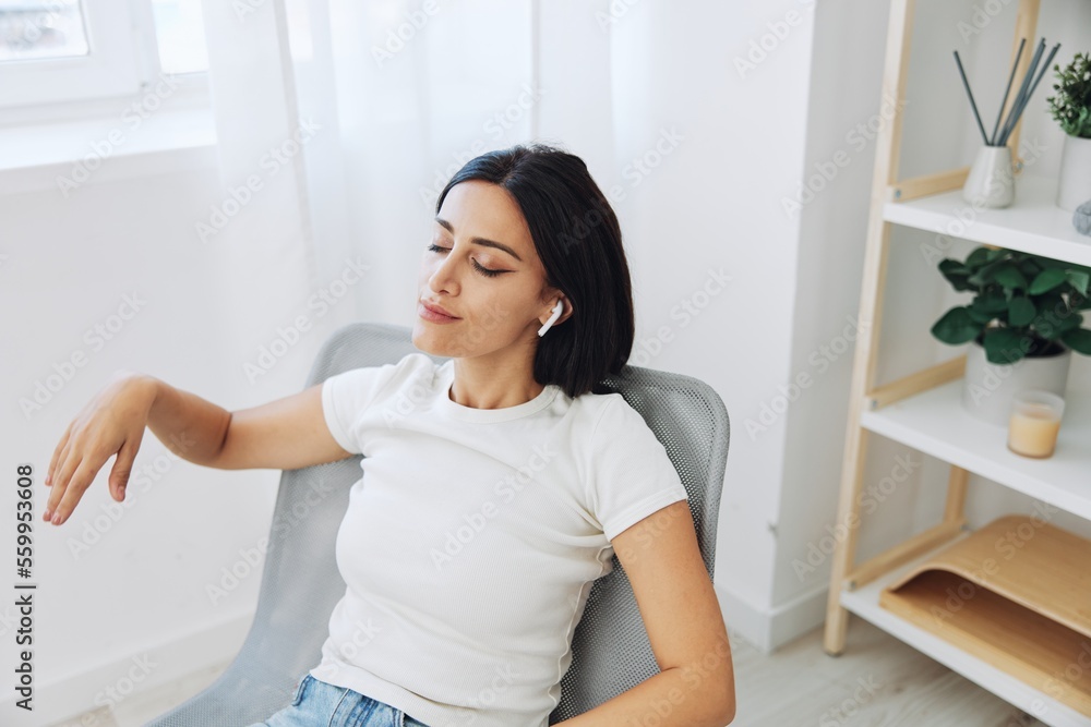 Woman sitting in a chair listening to music on wireless headphones at home in jeans and a white T-shirt, fall lifestyle comfort