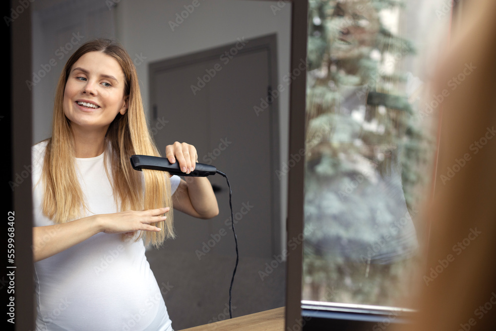 Fototapeta premium Pregnant woman straightening hair at bathroom using electric straightener iron. Beautiful lady taking care of hair 