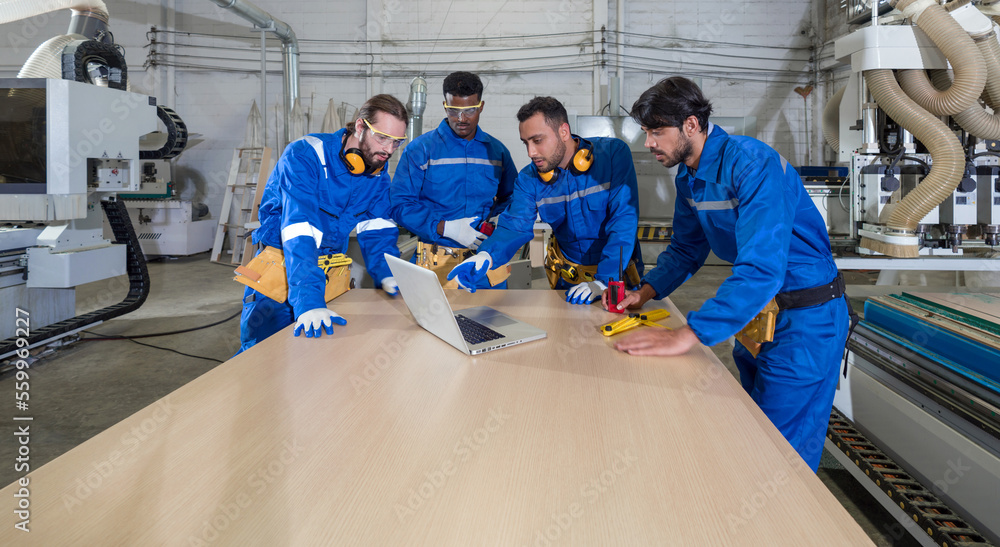 Obraz premium Group of multiethnic technicians in blue mechanic jumpsuits meeting, using laptop computers on large wooden table. Production line atmosphere in furniture factory