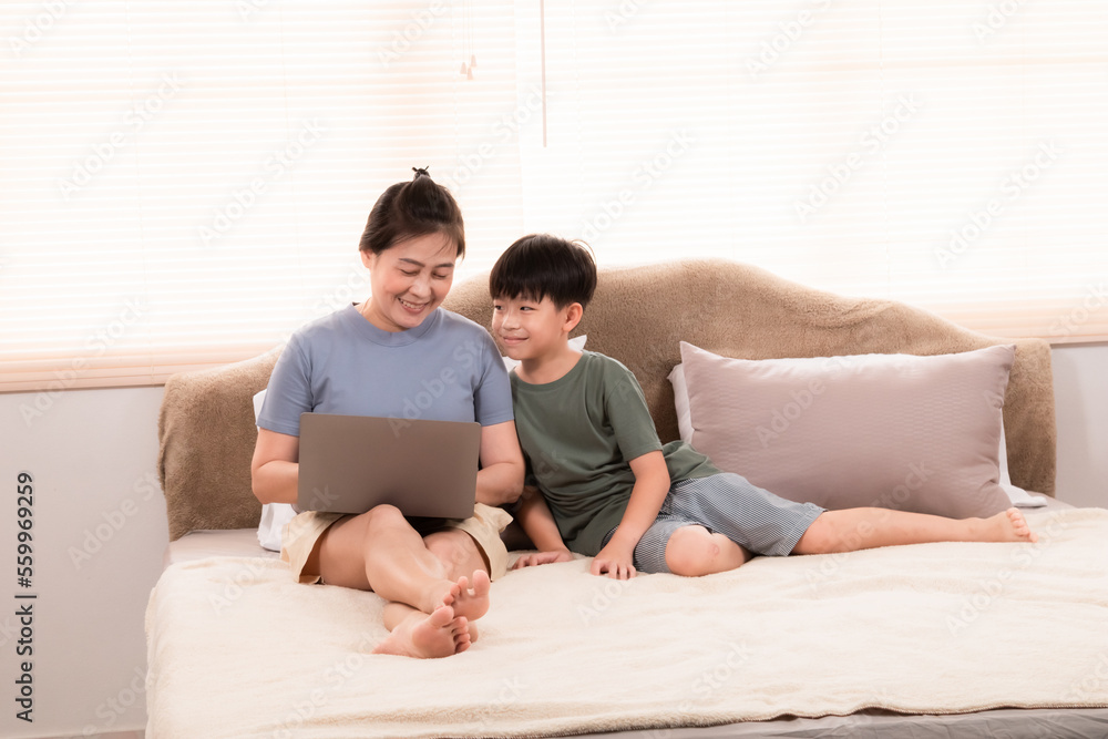 Asian happy mother using laptop working from home on bed while son sitting next to want to play with mom, Single mom work while raising boy. kid begging her for attention. Asian happy mother using laptop working from home on bed while son sitting next to want to play with mom, Single mom work while raising boy. kid begging her for attention.