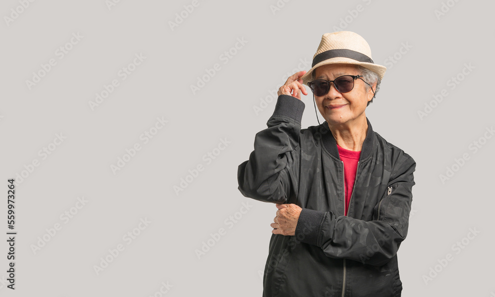 Senior woman in a black jacket wearing sunglasses and a hat looks at the camera with a smile while standing on a gray background Senior woman in a black jacket wearing sunglasses and a hat looks at the camera with a smile while standing on a gray background