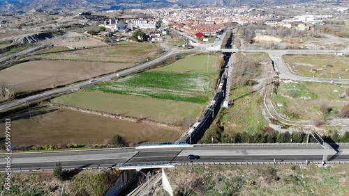 Aerial: regional train passing under a main road and entering a medium-sized town in Catalonia with the Pyrenees mountain range in the background photo