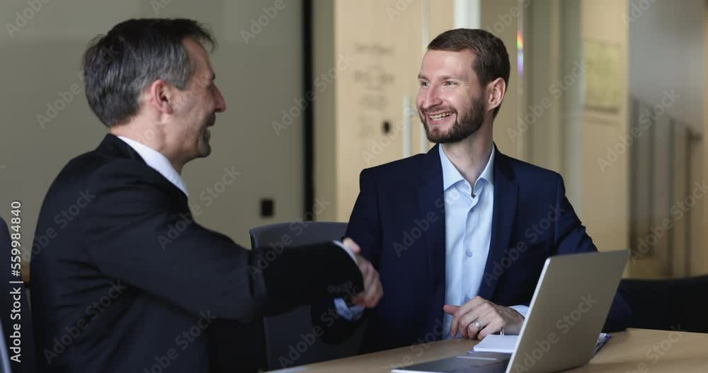 Two businessmen in suits sit at desk shake hands, finish formal meeting. Client and sales manager make deal, express respect handshake, feel satisfied by successful effective negotiations in office Two businessmen in suits sit at desk shake hands, finish formal meeting. Client and sales manager make deal, express respect handshake, feel satisfied by successful effective negotiations in office
